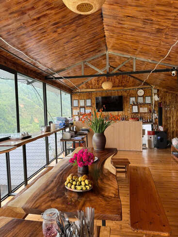 A cozy, wooden interior café with large windows overlooking green hills. Natural wood tables, flowers, and fruit create a warm, inviting atmosphere perfect for relaxation.