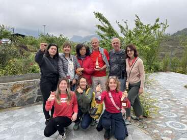 A imagem mostra um grupo de nove pessoas posando para uma foto, com oito mulheres e um homem, todos segurando garrafas de vinho ou champanhe, em um cenário externo.