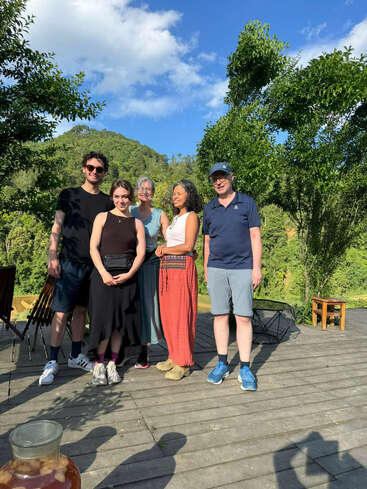 Cinco personas están juntas en una terraza de madera bañada por el sol, rodeadas de frondosos árboles y colinas, sonriendo y disfrutando de un hermoso día despejado al aire libre.