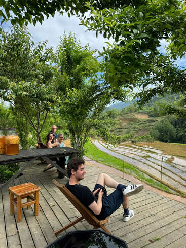 Cuatro personas descansan en sillas de madera en una terraza con vistas a unas pintorescas terrazas de arroz. Están rodeadas de frondosos árboles verdes. Unas jarras y una mesa de madera completan esta tranquila escena.