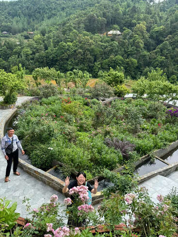 Dos personas disfrutan de un frondoso jardín en terrazas rodeado de una exuberante vegetación y flores en plena floración, con una ladera boscosa y casas dispersas como pintoresco telón de fondo.