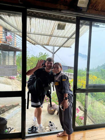 Um homem e uma mulher estão sorrindo em frente a uma porta de vidro, acenando. Flores coloridas, sapatos, vegetação e montanhas enevoadas criam um cenário ao ar livre tranquilo e acolhedor.