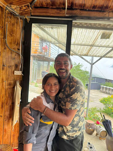 Um casal sorridente se abraça alegremente dentro de casa, perto de uma janela. Lá fora, há vegetação, vasos de plantas e um prédio com flores na varanda.