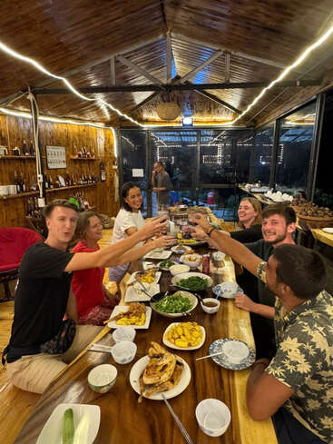 Um grupo de amigos se reúne em torno de uma mesa de madeira, compartilhando uma refeição e brindando em comemoração, dentro de um restaurante aconchegante, com iluminação suave e um ambiente rústico.