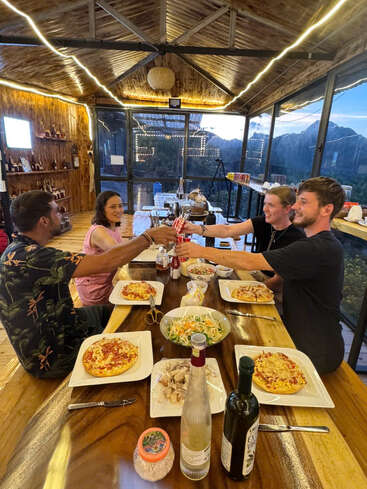Quatro pessoas estão jantando juntas numa cabana de madeira aconchegante, compartilhando pizza e bebidas, cercadas por montanhas e luzes aconchegantes, sorrindo e brindando alegremente.