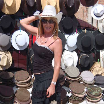 Une femme se tient devant un mur de chapeaux, vêtue d'un chapeau de cow-boy blanc et d'un débardeur noir, posant pour l'appareil photo, la main droite sur la tête.