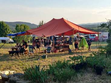 A group of people gather under a large red tent outdoors, surrounded by greenery. Tables, chairs, and camping equipment create a relaxed, communal, festival-like atmosphere.