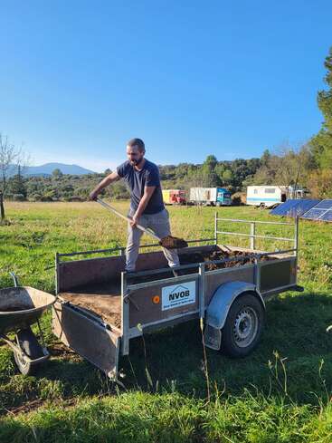 A man stands on a trailer filled with soil, shoveling dirt. He’s outdoors in a grassy field, with solar panels, trees, and vehicles in the background.