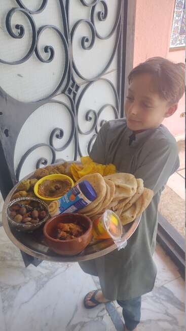Ein Junge, der traditionell gekleidet ist, hält ein großes Tablett mit verschiedenen Speisen wie Brot, Bohnen, Falafel, Curry, Joghurt und Nachtisch in der Hand und steht neben einer verschnörkelten Tür.