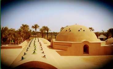 Dieses Bild zeigt einen traditionellen Kuppelbau aus Lehm mit bunten Glasakzenten, umgeben von Palmen unter einem hellen Himmel, der an eine Oase oder ein Wüstendorf erinnert.