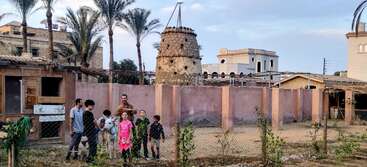 Eine Gruppe von Kindern und ein Erwachsener stehen mit einem Esel in einem eingezäunten Bereich, umgeben von Palmen, traditionellen Gebäuden und einem markanten Taubenturm.