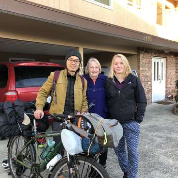 L'image montre trois personnes debout devant une maison avec un abri pour voiture, où un véhicule rouge est garé, et un vélo chargé de sacs est positionné devant eux.