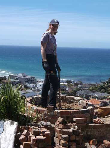 Una persona con guantes y sombrero está de pie sobre una estructura circular de ladrillo, con vistas a una pintoresca ciudad costera y a un vasto océano azul bajo un cielo despejado.
