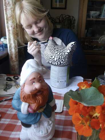 Una mujer sonriente pinta una figura decorativa de un pollo en una mesa con una estatua de un gnomo, flores naranjas y material de arte en una acogedora habitación iluminada por el sol.
