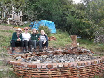 Cuatro personas se sientan juntas en un parterre de ladrillos lleno de piedras en un jardín. Detrás de ellos hay vegetación, una lona azul y estructuras de madera.