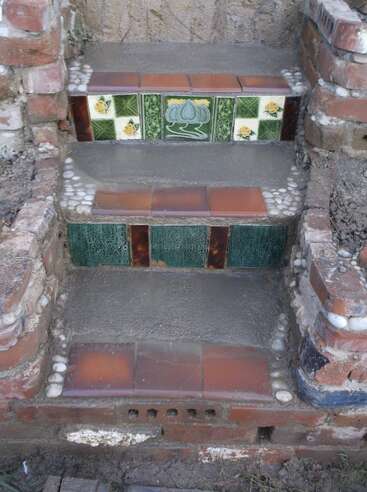 La imagen muestra unos escalones rústicos de ladrillo adornados con azulejos decorativos de cerámica, que incluyen motivos verdes, florales y de loto, con detalles de guijarros blancos incrustados en el hormigón.