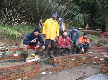 Cinco personas, embarradas y vestidas con ropa de trabajo, posan sobre un muro de ladrillos parcialmente construido y rodeado de vegetación, aparentando estar contentas y enfrascadas en un proyecto de construcción al aire libre.
