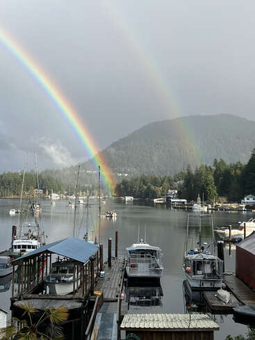 A beautiful double rainbow arches over a serene marina, with boats docked and calm water reflecting the vibrant colors, surrounded by lush green forested mountains.