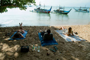 Três pessoas relaxam em toalhas sob uma árvore, observando barcos ancorados em águas calmas. Chinelos, bebidas e sombra realçam a cena tranquila da praia.