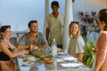 Um grupo de pessoas está sentado ao redor de uma mesa ao ar livre, desfrutando de uma refeição. Elas estão sorrindo, conversando e cercadas por pratos e bebidas.