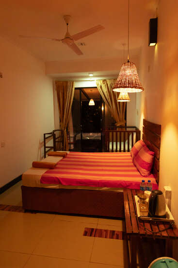 Um quarto aconchegante e bem iluminado com uma cama listrada, luminárias pendentes de vime, cabeceira de madeira, varanda, cortina, ventilador de teto e uma mesa lateral com garrafas de água e uma chaleira.