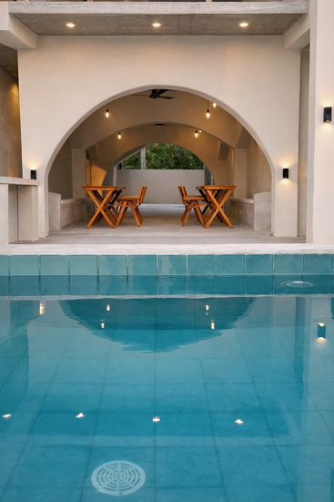 Una piscina serena refleja azulejos turquesa y un moderno patio abovedado con mesas de madera, luces cálidas y un diseño arquitectónico tranquilo y minimalista en tonos suaves.