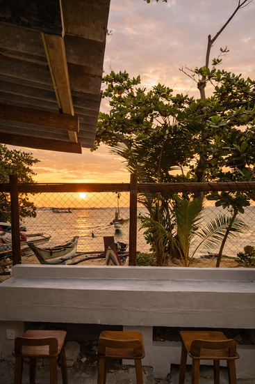 Taburetes de madera frente a una barra de hormigón con vistas a una playa tropical al atardecer. Barcos flotan en aguas tranquilas, enmarcados por frondosos árboles verdes y un cielo cálido.