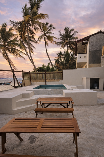 Dos mesas de madera se encuentran en un patio de hormigón frente a una pequeña piscina, rodeadas de palmeras, una valla y una casa al atardecer. Ambiente tranquilo.