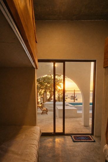 La suave luz dorada del sol se filtra a través de las puertas de cristal, iluminando una acogedora habitación con literas. En el exterior, un patio abovedado da a una tranquila piscina y al sereno océano al atardecer.