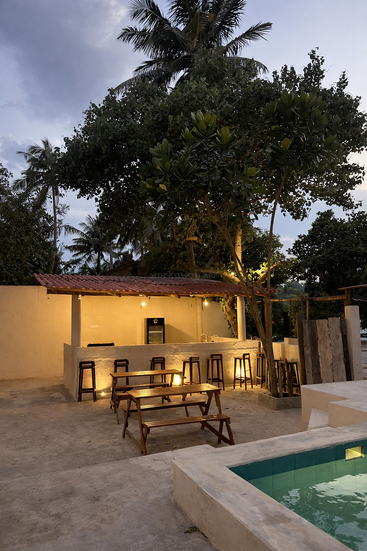 Una zona de bar al aire libre con bancos y taburetes de madera, iluminada por luces cálidas, se encuentra cerca de una pequeña piscina. Altos árboles tropicales y el cielo del atardecer completan la escena.
