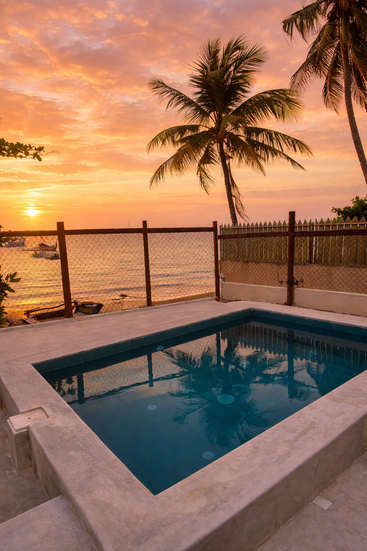 Una tranquila escena costera al atardecer, con una pequeña piscina, palmeras y olas tranquilas, con tonos dorados y rosados reflejándose en el agua y el cielo.