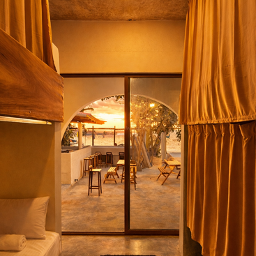 Une lumière dorée et chaleureuse emplit une chambre d'auberge confortable équipée de lits superposés et de rideaux, donnant sur un patio et un coucher de soleil paisible sur la plage à travers des portes vitrées.