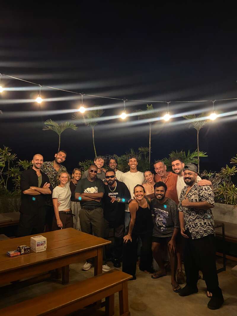 Um grupo de quinze pessoas posa junto ao ar livre à noite, sob luzes de cordas, cercado por plantas tropicais. Todos estão sorrindo, criando uma atmosfera alegre e amigável.