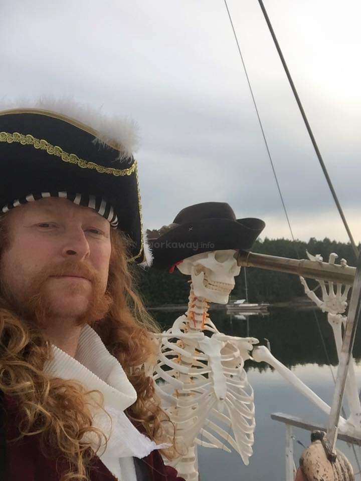 L'image représente un homme habillé en pirate à côté d'un squelette portant un chapeau, avec un plan d'eau et des arbres en arrière-plan par temps couvert.
