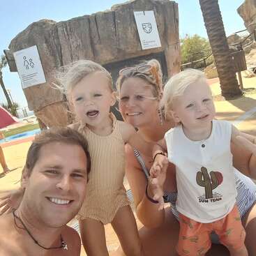 Eine lächelnde vierköpfige Familie posiert im Freien unter der Sonne. Zwei Erwachsene und zwei kleine Kinder genießen einen sonnigen Tag an einem Pool mit Palmen.