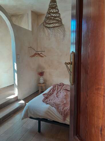 Dieses gemütliche, minimalistische Schlafzimmer zeichnet sich durch ein einfaches Bett mit einer rosa Decke, natürliche Beleuchtung, eine rustikale Lampe, eine dekorative Vase, einen Holzhocker, Wandaufhänger und warme Töne aus.