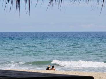 Um casal está sentado na areia da praia, perto das ondas suaves, sob um céu azul claro, com folhas de palmeira emoldurando a parte superior da cena tranquila.