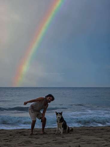Um homem e um cachorro husky posam em uma praia de areia. Atrás deles, ondas vibrantes batem e um arco-íris vívido se estende lindamente pelo céu enevoado.