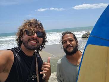 Dois homens posam para uma selfie em uma praia ensolarada. Um deles segura um sinal de shaka, de óculos escuros, com uma prancha de surfe visível e as ondas do mar atrás deles.