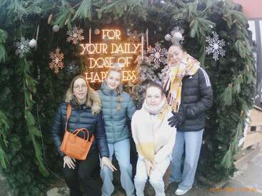 Vier Frauen in Winterkleidung posieren vor einer festlichen Kulisse aus Tannen und Schneeflocken. Auf einem leuchtenden Schild steht: "Für deine tägliche Dosis Glück".