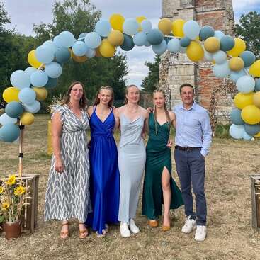 A family of five poses under a decorative balloon arch outdoors. The women wear elegant dresses, the man is in shirt and pants. Sunflowers decorate the scene.