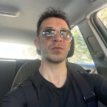 L'image montre un homme aux cheveux noirs et aux lunettes de soleil prenant un selfie sur le siège avant d'une voiture, portant une chemise noire et une ceinture de sécurité.