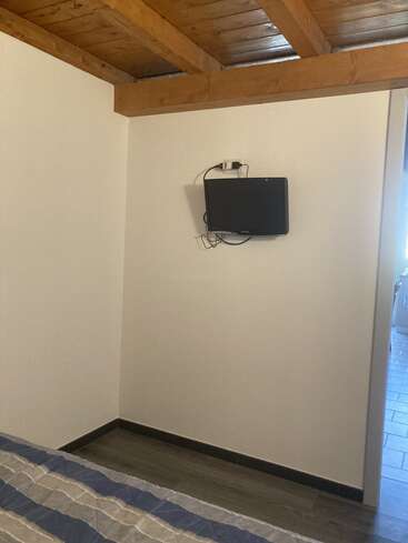L'image représente une chambre aux murs blancs, au plafond en bois, avec un téléviseur fixé au mur, un lit et une porte menant à une autre pièce.