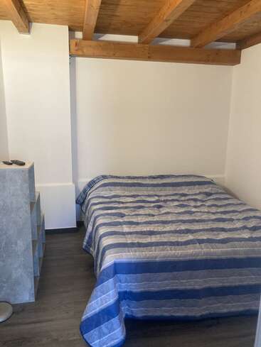 L'image représente une petite chambre confortable avec un lit simple, des poutres en bois au plafond et une commode, avec des murs blancs et un sol en bois foncé.