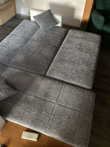 L'image représente un canapé gris avec une chaise longue, accompagné d'un petit coussin gris, situé dans un salon aux murs blancs et au sol en bois foncé.