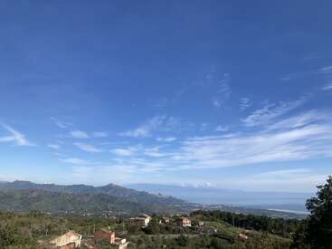 L'image représente un paysage serein avec une petite ville nichée au milieu de collines verdoyantes, avec une vaste étendue de ciel bleu au-dessus et une étendue d'eau lointaine.