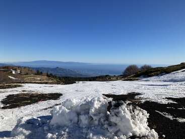 L'image représente un paysage serein et enneigé, avec une vaste étendue de ciel bleu, une chaîne de montagnes lointaine et un petit groupe d'arbres au premier plan.