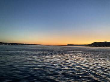 L'image représente une scène de lac sereine au coucher du soleil, avec une vaste étendue d'eau et une chaîne de montagnes lointaine sous un ciel dégradé de teintes bleues et orange.