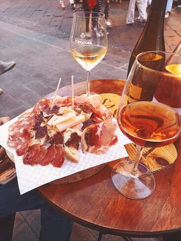 L'image représente une table en bois avec une planche de charcuterie, deux verres de vin et une bouteille de vin, sur fond de trottoir avec des gens qui marchent.
