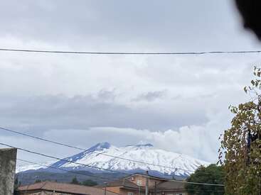 L'image représente une montagne enneigée sous un ciel nuageux, avec des lignes électriques et des bâtiments au premier plan, et des arbres sur le côté droit.